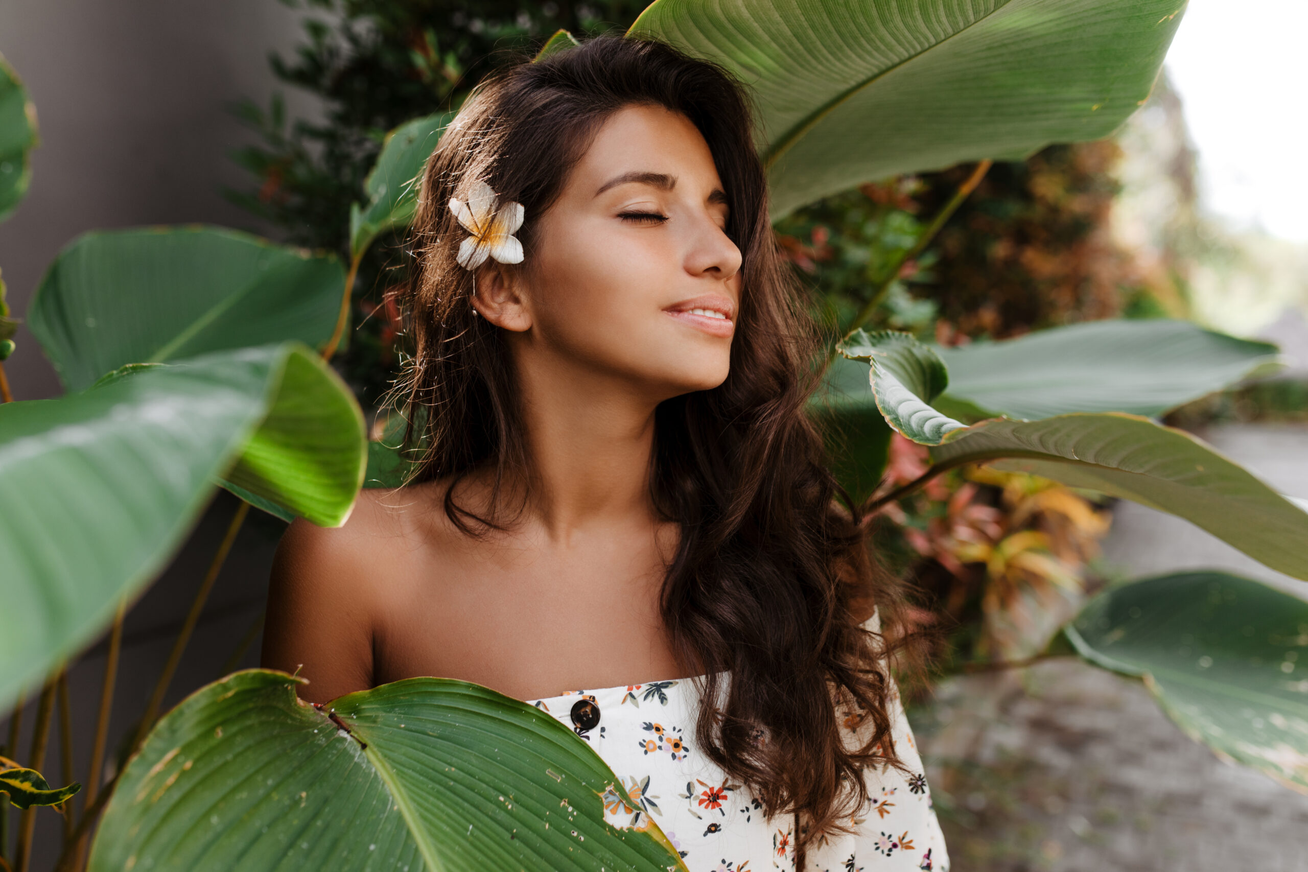 Close-up portrait of pacified woman with flower in dark wavy hair. Lady with slight smile and closed eyes posing against backdrop of tropical plant